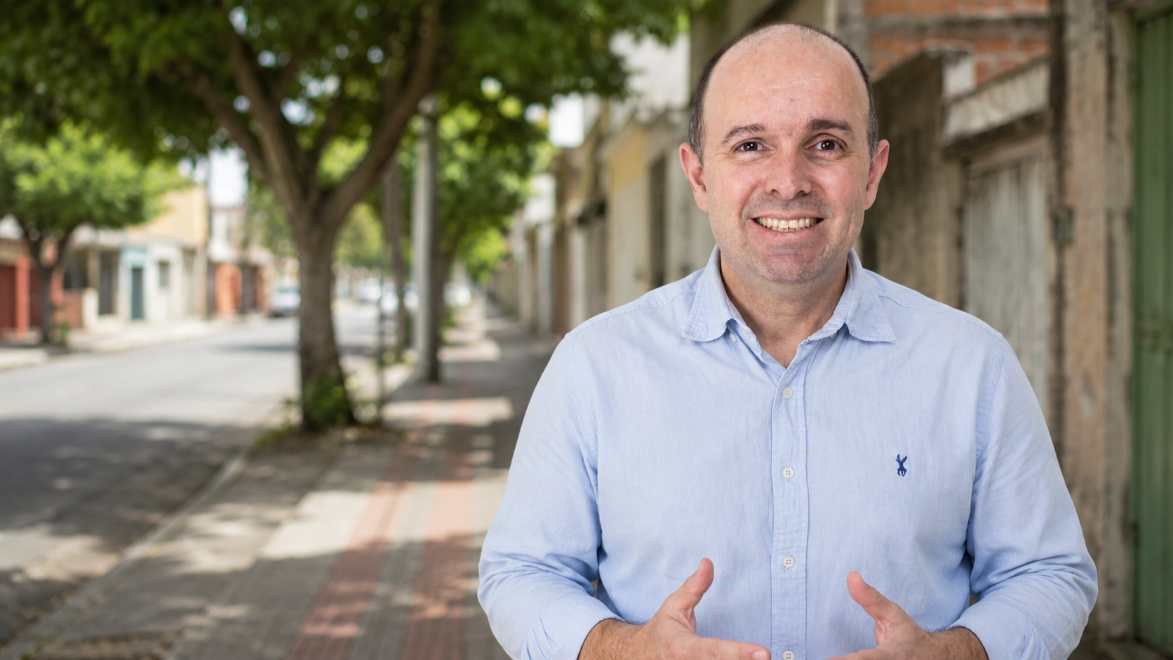 Angelo Cesário, de camisa azul-clara, conversando em uma rua arborizada de bairro.